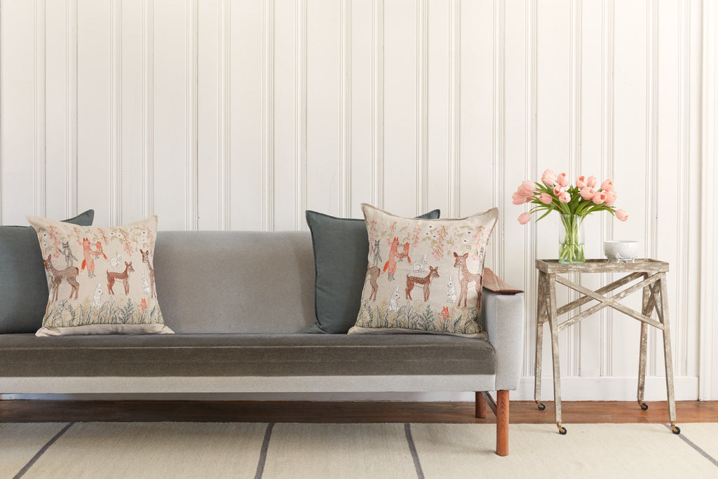 A cozy living room corner featuring a gray sofa with a Coral & Tusk Spring Blossoms Pillow and decorative cushions, a small side table with a vase of pink tulips, against a white paneled wall.