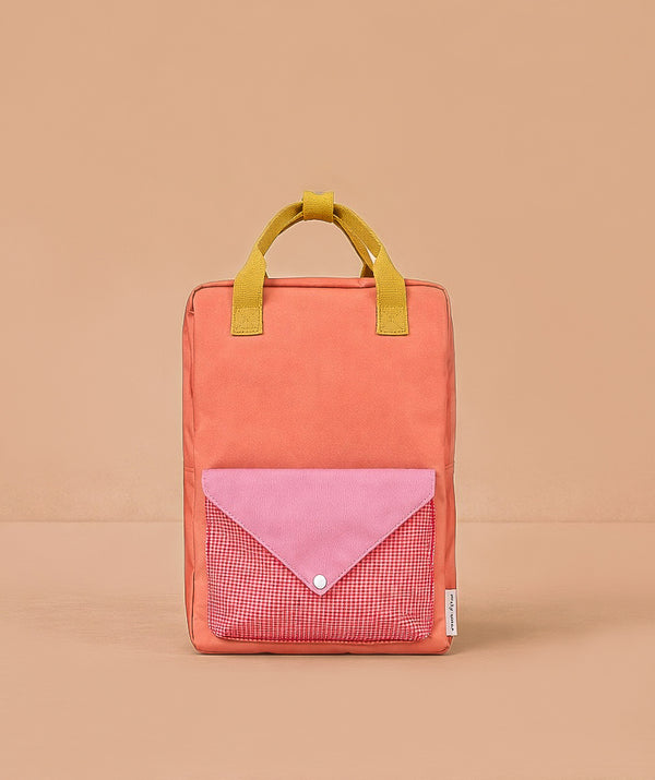 The Sticky Lemon Backpack Large - Envelope - Maple Whirl is a rectangular orange kids’ backpack made from recycled PET bottles, with yellow handles and a front pocket featuring a pink flap and red gingham, shown on a light brown background.