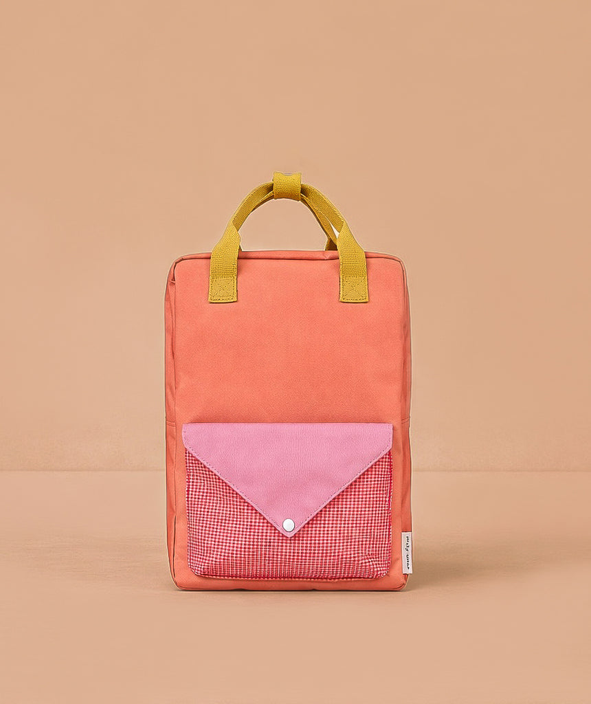The Sticky Lemon Backpack Large - Envelope - Maple Whirl is a rectangular orange kids’ backpack made from recycled PET bottles, with yellow handles and a front pocket featuring a pink flap and red gingham, shown on a light brown background.