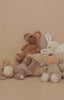 Four plush toys with closed eyes, dressed as a bear, bunny, and two lambs, are arranged on a beige background beside a textured cushion. Spot the cuddly Olli Ella Cozy Dinkum Doll – Lamby Pip among them.