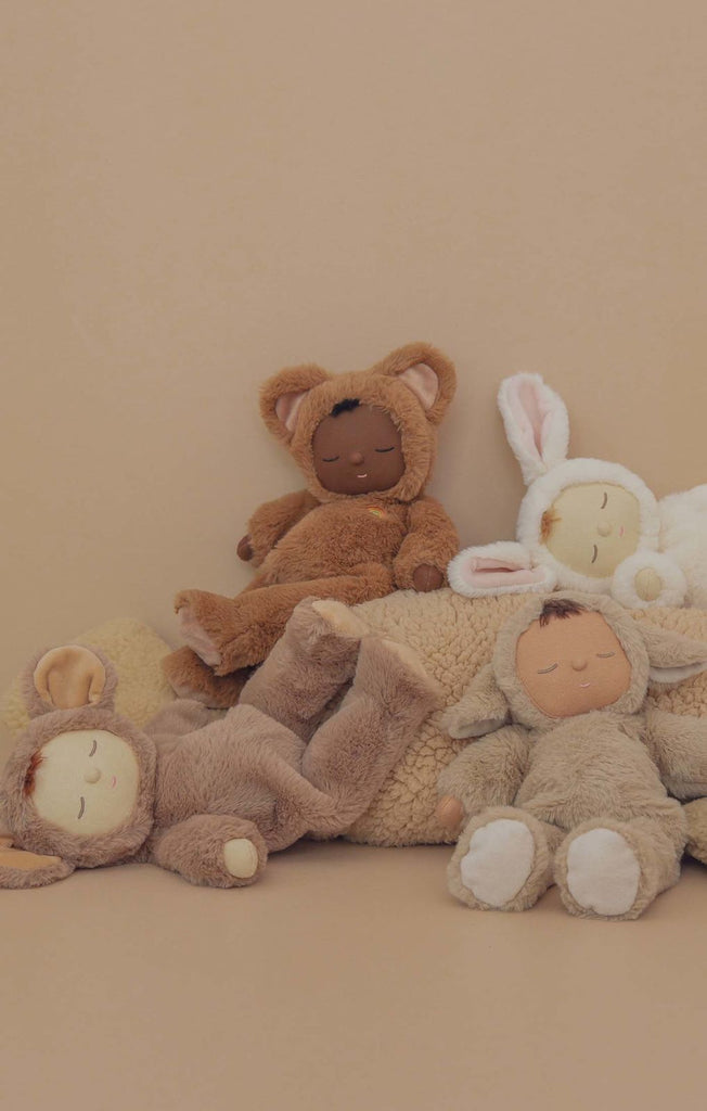 Four plush toys with closed eyes, dressed as a bear, bunny, and two lambs, are arranged on a beige background beside a textured cushion. Spot the cuddly Olli Ella Cozy Dinkum Doll – Lamby Pip among them.