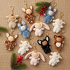 Twelve Olli Ella Cozy Bag Charms—Lamby Pip, shaped like sleeping babies in lamb costumes—are arranged on burlap with pine and berry sprigs. These plush keychains make adorable accessories for bags or keys.