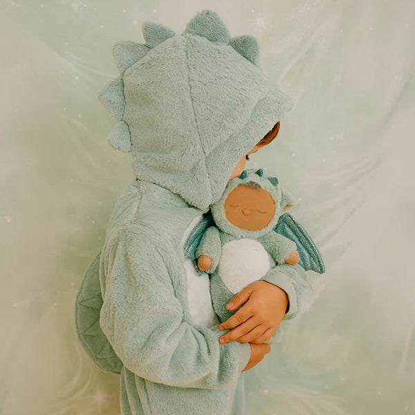 A child in a light blue dinosaur costume hugs the Olli Ella Cozy Dinkum Dragon - Ash - Green, a recycled plush toy with small wings and closed eyes, against a softly blurred background.