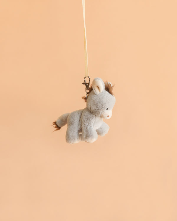 Bukowski’s Christopher Donkey Bag Charm, a small gray plush donkey toy, hangs by a thin strap and metal clip against a solid peach-colored background—perfect as a whimsical keychain or bag accessory.