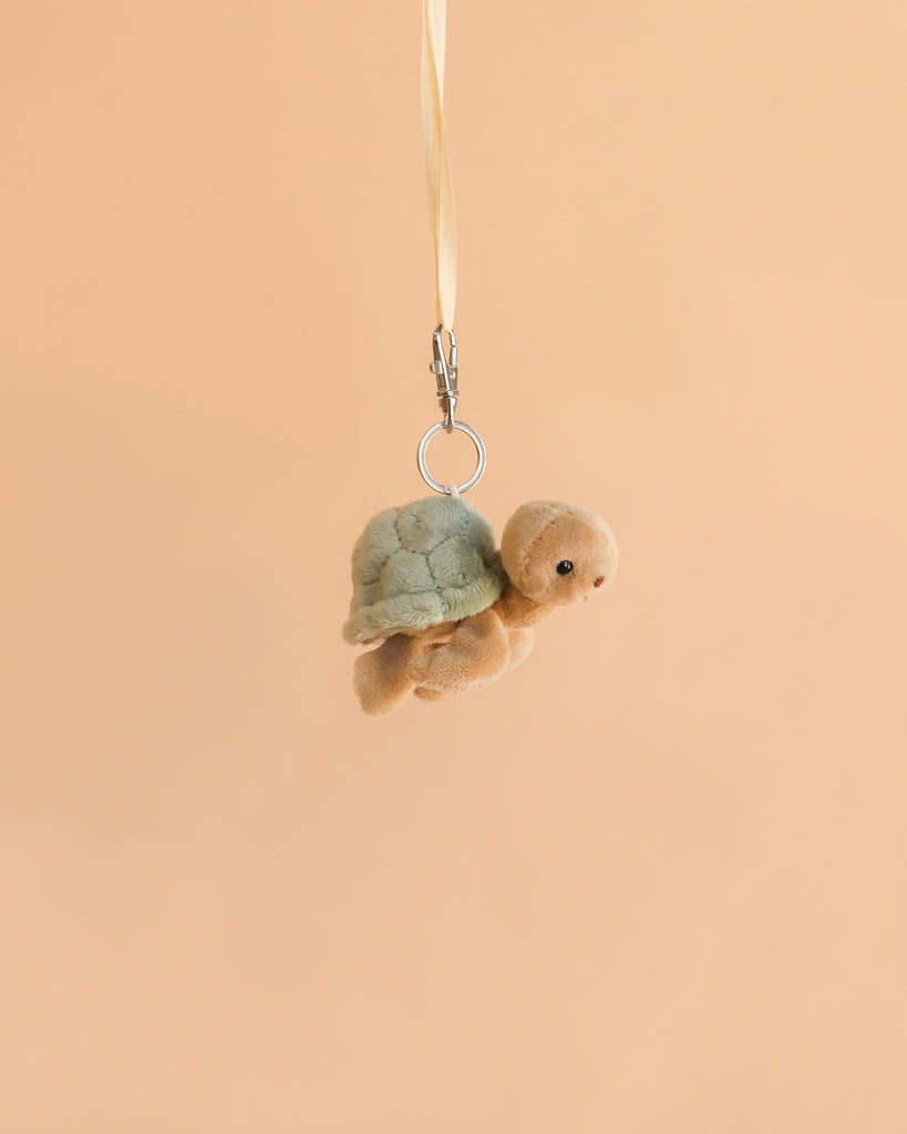 The Bukowski - Sandy Green Turtle Bag Charm features a soft plush sea turtle with a green shell, hanging from a yellow ribbon against a plain beige background, making it an adorable accessory for your bag.