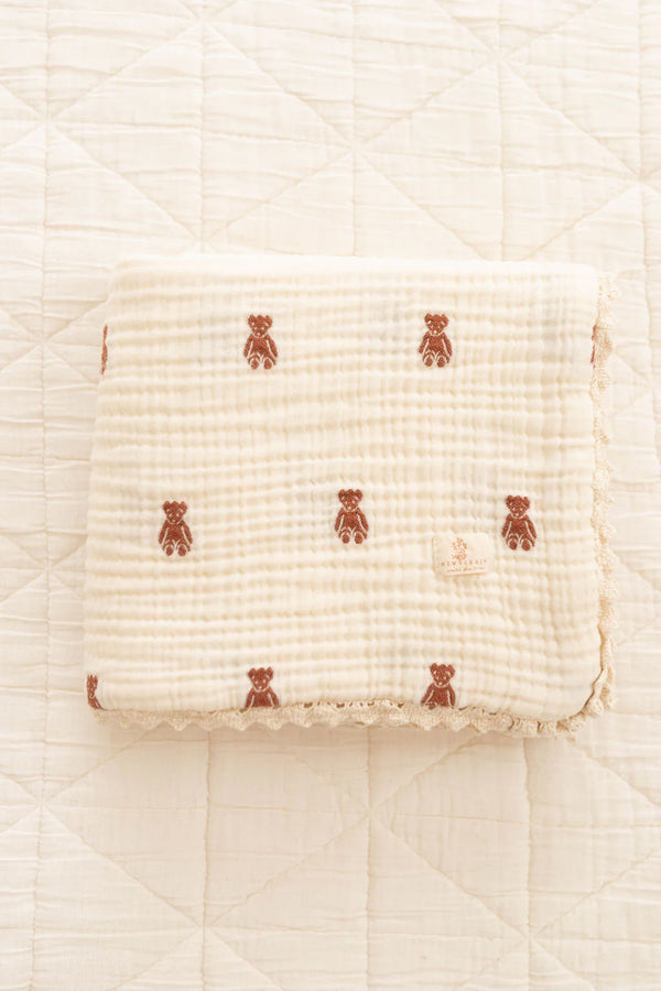 The Organic Cotton Layered Gauze Baby Blanket - Teddy (ships in one week), a soft, cream-colored blanket with a quilted texture and brown teddy bear patterns, lies neatly folded on a matching quilted surface.