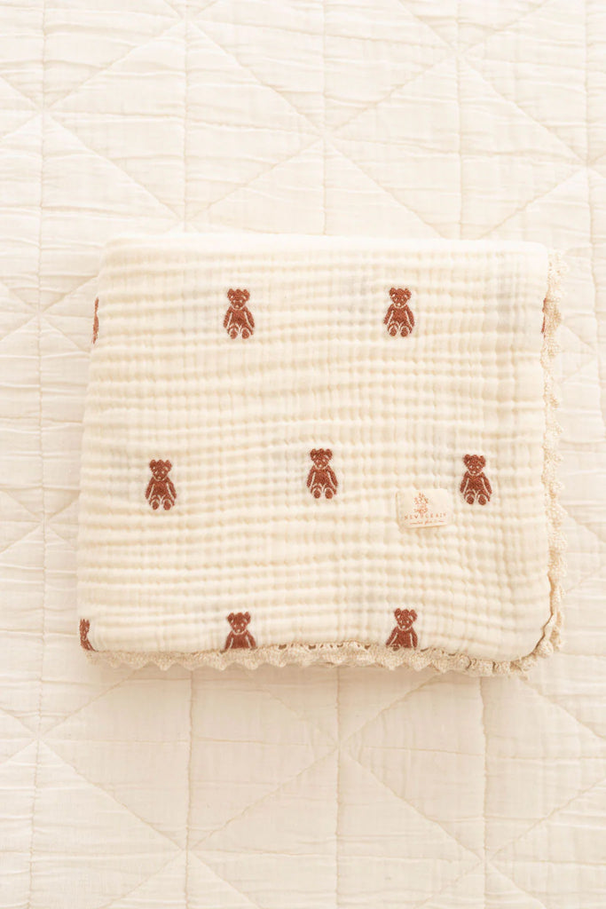 The Organic Cotton Layered Gauze Baby Blanket - Teddy (ships in one week), a soft, cream-colored blanket with a quilted texture and brown teddy bear patterns, lies neatly folded on a matching quilted surface.