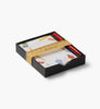 A black gift box contains the Schoolhouse Note Writing Set, featuring a notepad decorated with colorful doodles and two red pens. The box is wrapped with a gold paper band designed like a ruler and labeled “Desk Set,” making it an ideal addition to any reusable desk organizer.