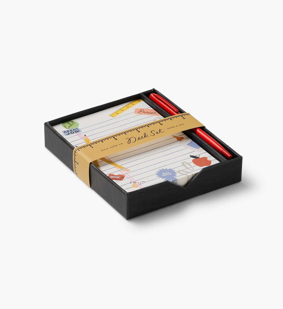 A black gift box contains the Schoolhouse Note Writing Set, featuring a notepad decorated with colorful doodles and two red pens. The box is wrapped with a gold paper band designed like a ruler and labeled “Desk Set,” making it an ideal addition to any reusable desk organizer.