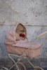 The Konges Sloejd Doll Pram - Cherry Blush sits against a textured concrete wall, featuring a vintage style with a soft pink canopy and quilted fabric. The pram is equipped with a padded mattress, handlebar, and two dangling red and brown toys above the bassinet.