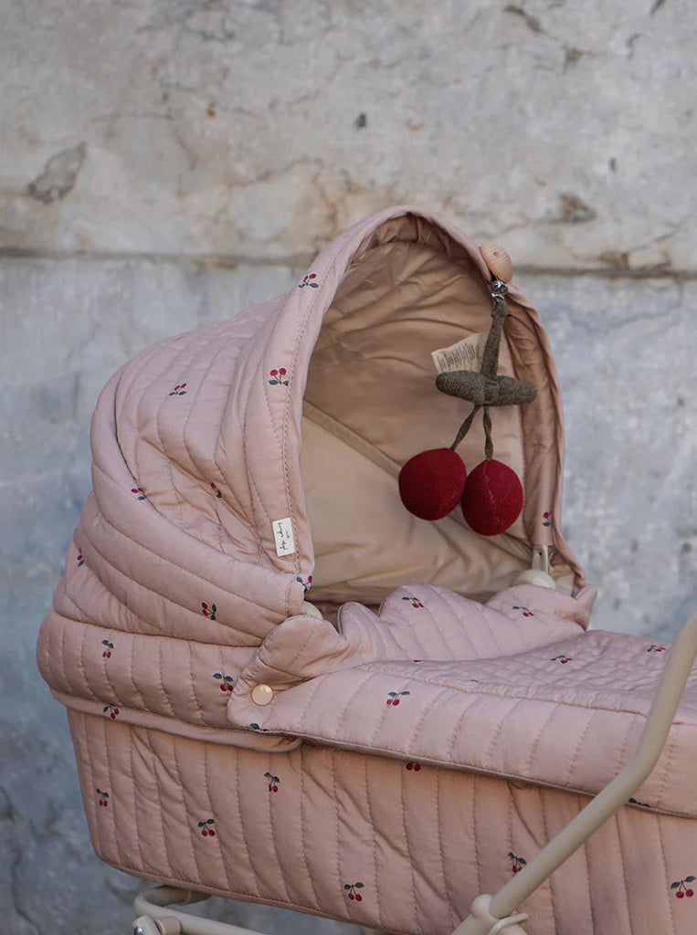 The Konges Sloejd Doll Pram - Cherry Blush embodies a vintage style with its pink quilted fabric cover adorned with small cherry patterns. Inside, it includes a toy set featuring a brush and two cherry-shaped plush toys. The textured gray wall in the background enhances its charming aesthetic.