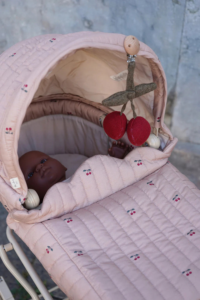 A baby rests in the Konges Sloejd Doll Pram - Cherry Blush, showcasing a quilted fabric cover adorned with small cherry motifs and a hanging toy with red cherries above. The scene is set against a blurred outdoor backdrop.