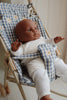 A baby doll with brown skin is seated in a Konges Sloejd Doll Stroller - Blue Checkered adorned with small white flowers. The stroller features double wheels for easy maneuverability. The doll, dressed in a white knitted outfit, appears to be looking upwards with its arms slightly raised. The background is softly lit and neutral in color.
