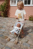 A young child with blonde hair is standing outside on a cobblestone path, pushing a Konges Sloejd Doll Stroller - Blue Checkered with double wheels and removable fabric. The child is wearing a beige shirt with a bear graphic and striped pants. A pink building with white-framed windows and green plants is in the background.