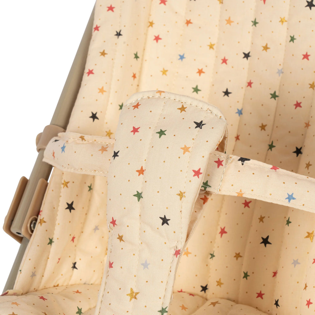 A close-up of a padded fabric seat cover in beige, adorned with colorful star patterns in red, green, blue, yellow, and black. The quilted stitching and the strap with matching star designs suggest it belongs to a Konges Sloejd Doll Stroller - Multi Star.