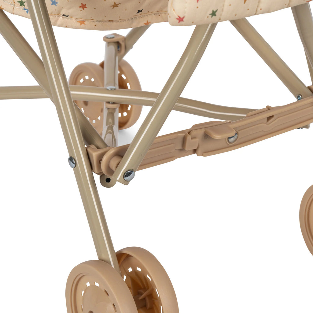 Close-up of a beige Konges Sloejd Doll Stroller - Multi Star's frame and double wheels. The fabric, adorned with small multicolored stars, complements the sturdy metal and plastic structure, showcasing the hinges, folding mechanism, and secure harness for added safety.