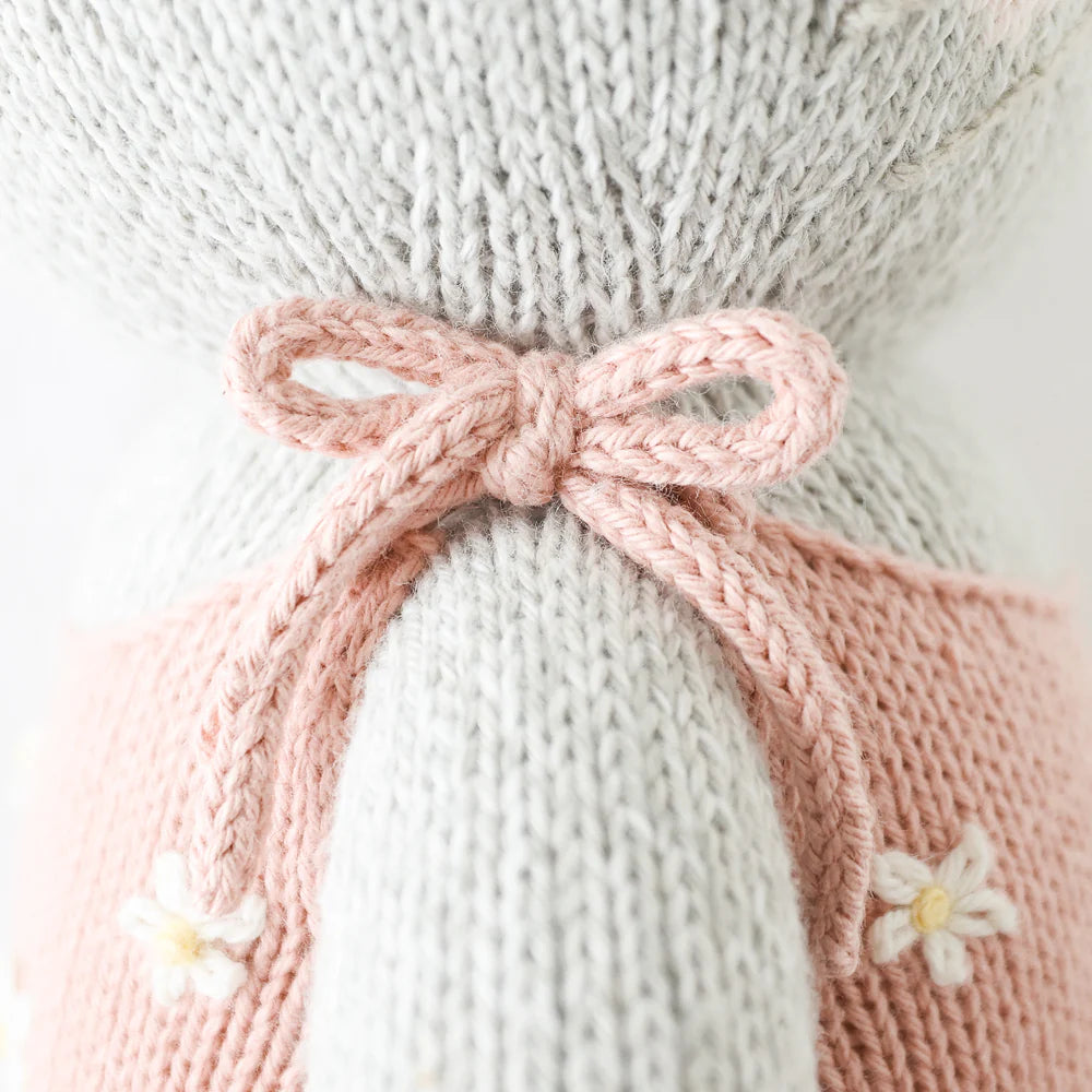 Close-up of Cuddle + Kind Daisy the Kitten, a hand-knit light gray animal doll in a pink dress with white embroidered flowers and a pink bow. Made from premium cotton yarn, it has a soft, textured finish.