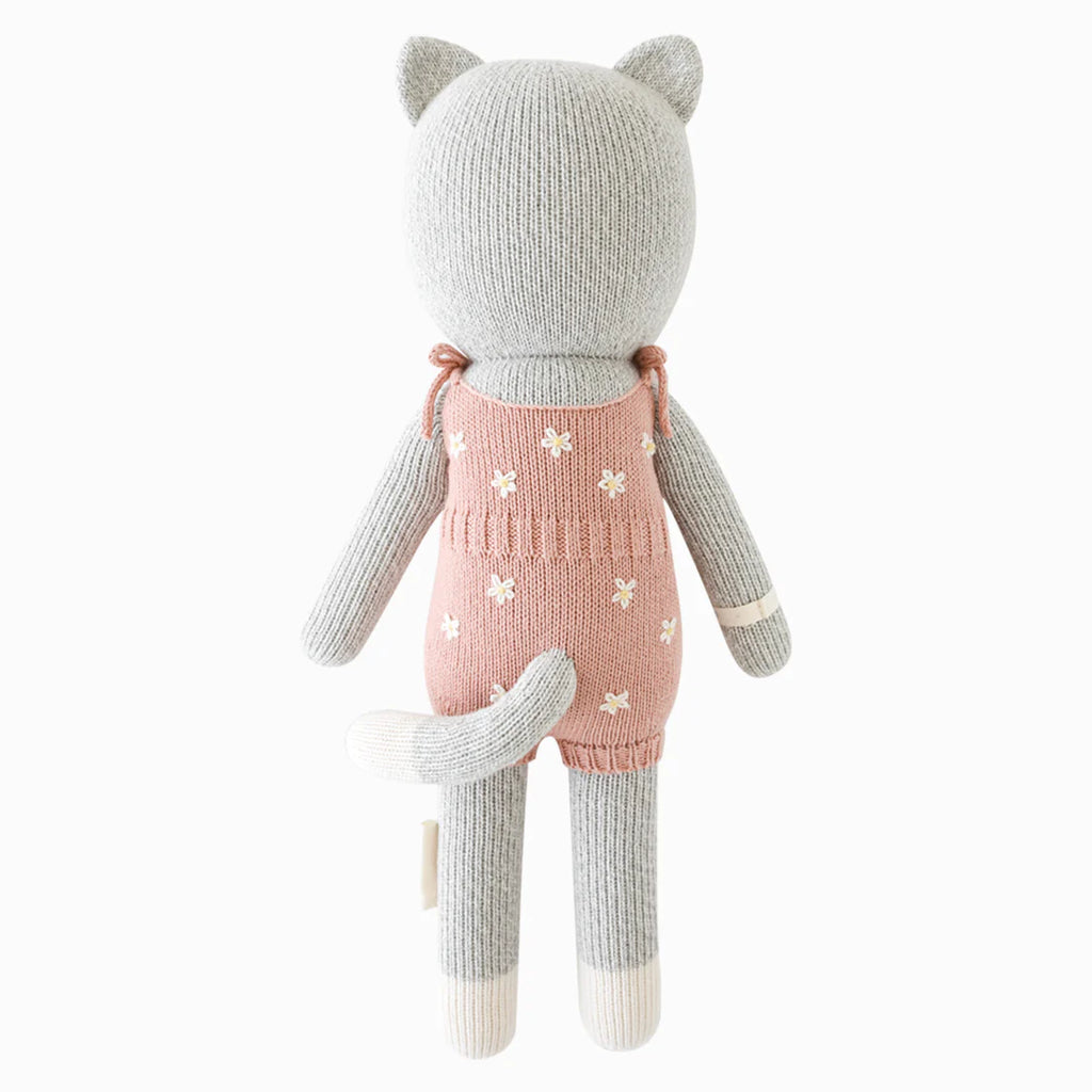 Cuddle + Kind Daisy the Kitten is a hand-knit, gray cotton doll in pink overalls with white stars. Seen from the back, it has a tail, cat ears, and cream accents on its paws and feet.