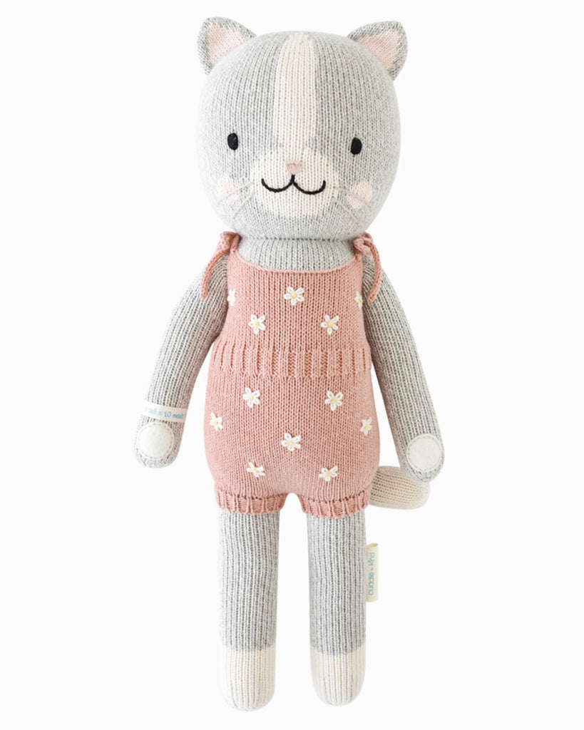Cuddle + Kind Daisy the Kitten is a hand-knit gray cat doll with a white snout, pink cheeks, and a pink romper with white flowers, crafted from premium cotton yarn and shown on a white background.