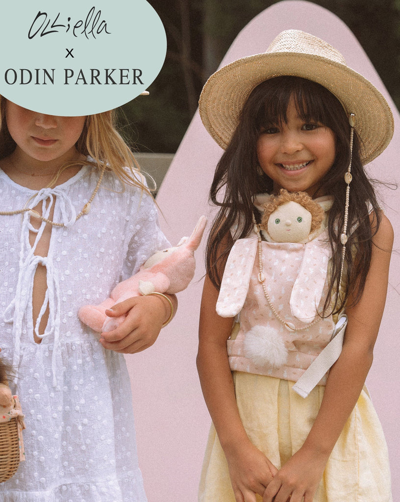 Two young girls smiling, one wearing a straw hat and holding a Dinkum Dolls Cottontail Carrier – Lapin in a kid-sized carrier, with a pink surfboard in the background. Text overlay says "Olli Ella x Odin Parker".