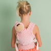 A young girl with blonde hair in a bun faces away, wearing a pink polka-dot dress and carrying the Olli Ella Petal Doll Carrier - Strawberry on her back, with her toy baby doll snugly tucked inside, against a light green background.