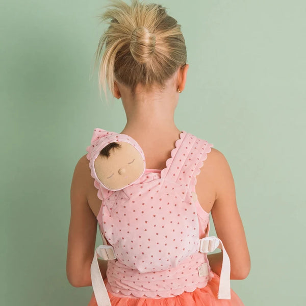 A young girl with blonde hair in a bun faces away, wearing a pink polka-dot dress and carrying the Olli Ella Petal Doll Carrier - Strawberry on her back, with her toy baby doll snugly tucked inside, against a light green background.