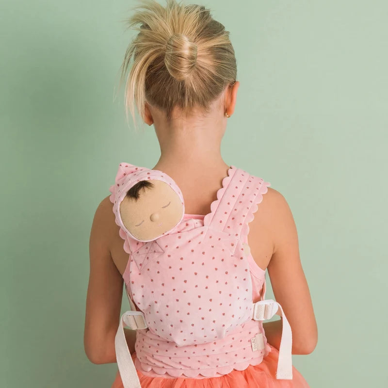 A young girl with blonde hair in a bun faces away, wearing a pink polka-dot dress and carrying the Olli Ella Petal Doll Carrier - Strawberry on her back, with her toy baby doll snugly tucked inside, against a light green background.