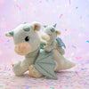 Two adorable Olli Ella Dinkum Dragon - Blaze plush toys in light green, one large and one small, with shiny wings. The smaller dragon rides on the larger one's back amid colorful confetti on a pastel background.