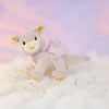 The Olli Ella Dinkum Dragon - Flare - Lavender is a plush lavender toy with shiny wings, resting on white clouds beneath a pastel pink and purple sky sparkling in the background.