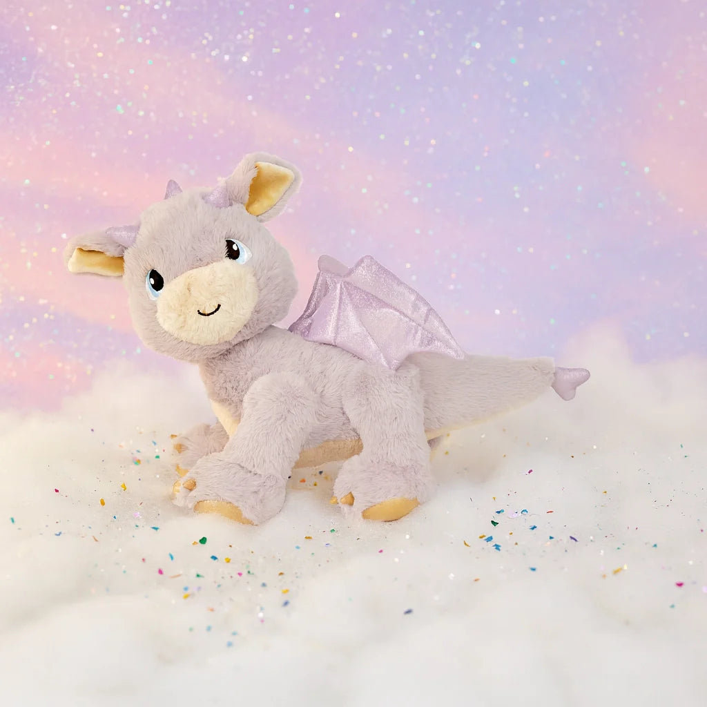The Olli Ella Dinkum Dragon - Flare - Lavender is a plush lavender toy with shiny wings, resting on white clouds beneath a pastel pink and purple sky sparkling in the background.