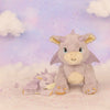 Two plush Olli Ella Dinkum Dragon - Flare - Lavender toys sit on a soft, pastel surface with dreamy clouds and a starry lavender sky—one upright and smiling, the other peacefully asleep, like magical companions in an enchanting world.