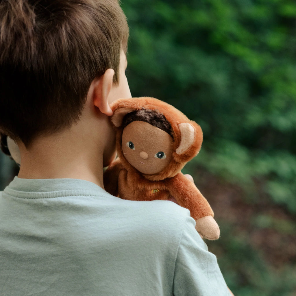 A young boy with his back to the camera holds a plush Olli Ella | Dinky Dinkums Forest Friends - Bobby Bear toy over his shoulder, looking ahead at a blurred green background.