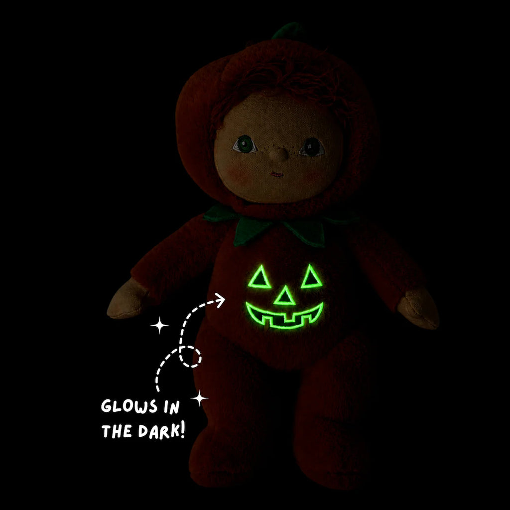 A dark-colored Olli Ella Dinky Dinkum Doll - Jules Jack-O’-Lantern plush in a pumpkin costume, with a glowing green jack-o’-lantern face on its chest and text that reads “Glows in the dark!” with an arrow pointing to the design.
