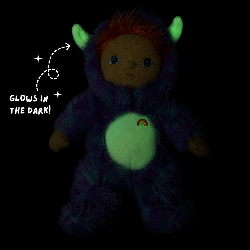 An Olli Ella Dinky Dinkum Doll - Miles Monster glow-in-the-dark plush toy with green horns and belly, wearing a fuzzy purple suit with a rainbow emblem, next to text that reads “Glows in the dark!”.
