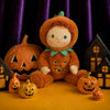 An Olli Ella Dinky Dinkum Doll - Jules Jack-O’-Lantern in a fuzzy orange pumpkin costume sits surrounded by small pumpkins and two dark house-shaped decorations.