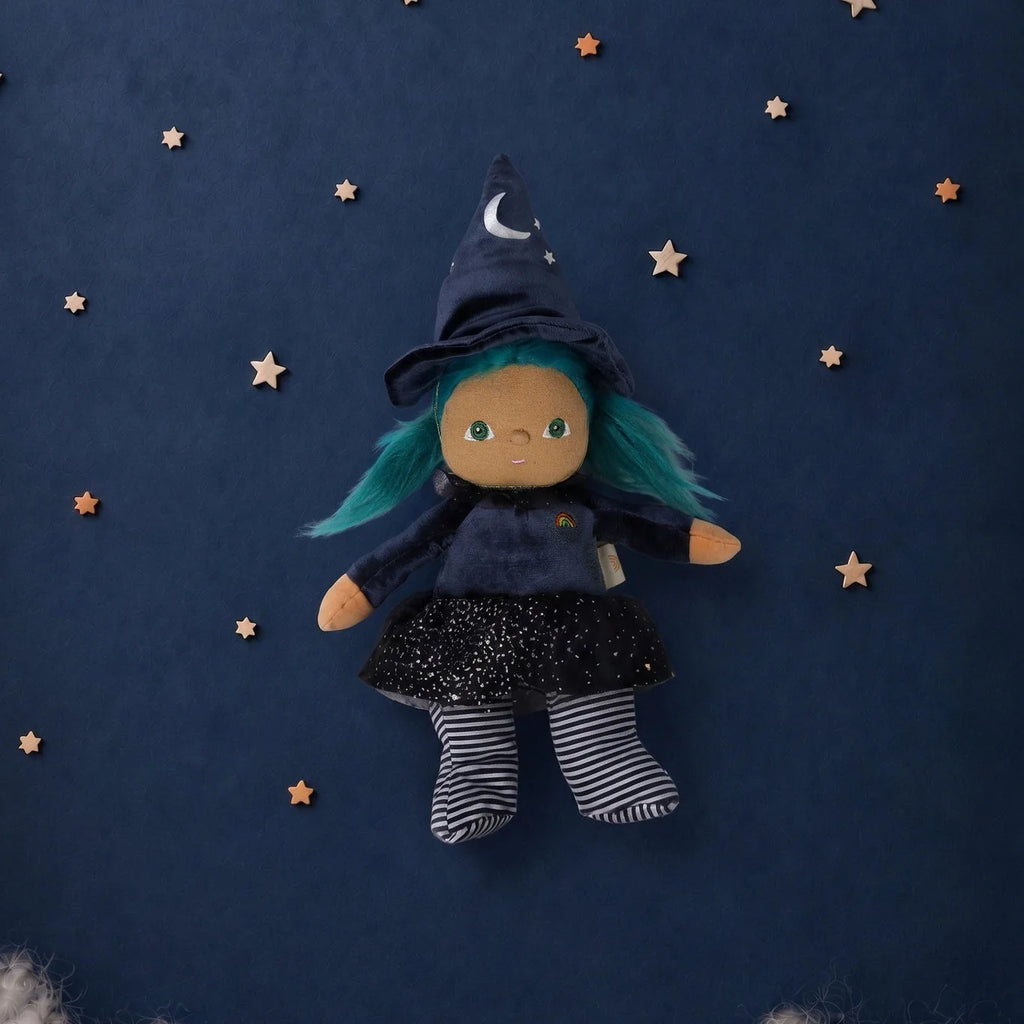 An Olli Ella Dinky Dinkum Doll - Winnie Witch dressed in a pointed hat, blue hair, and a starry skirt, surrounded by small wooden stars on a dark blue background.