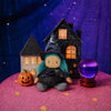 An Olli Ella Dinky Dinkum Doll - Winnie Witch dressed in a witch costume with teal hair, sitting between a miniature toy house, a carved pumpkin, and a glowing purple crystal ball on a pink table.
