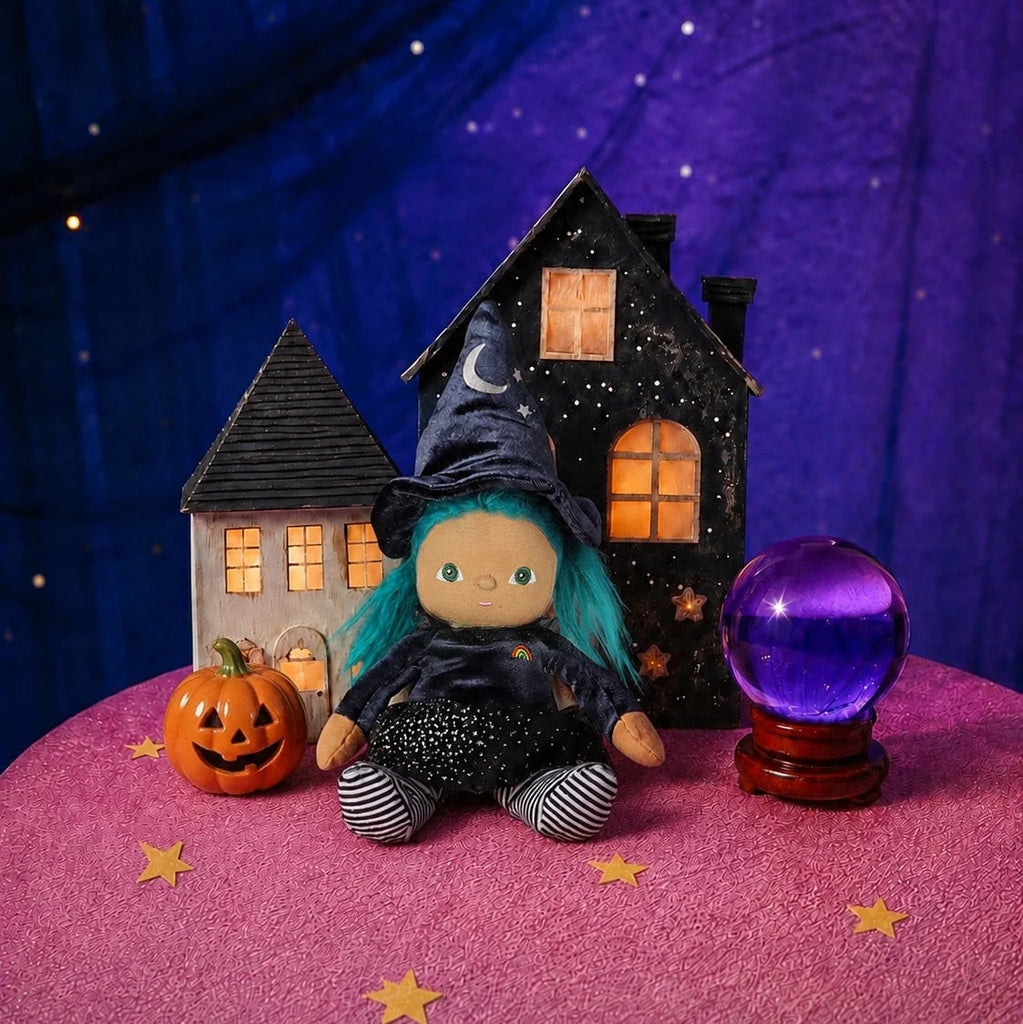 An Olli Ella Dinky Dinkum Doll - Winnie Witch dressed in a witch costume with teal hair, sitting between a miniature toy house, a carved pumpkin, and a glowing purple crystal ball on a pink table.