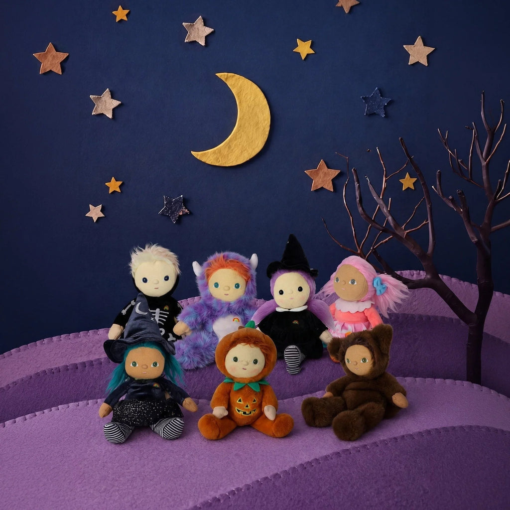 Seven dolls, including an Olli Ella Dinky Dinkum Doll - Winnie Witch, sit in various Halloween costumes on a purple hills landscape under a night sky with a moon and stars.