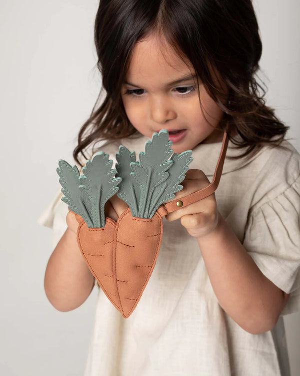 A young girl with shoulder-length brown hair examines a Donsje Chaeso Purse with a carrot design, holding it up against a light background.