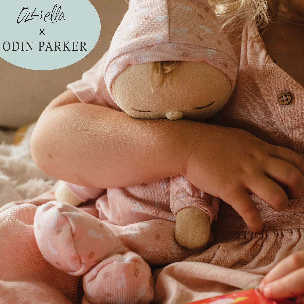 A young child hugging a soft Olli Ella x Odin Parker Dozy Dinkums - Blossom doll, dressed in a pink outfit with a patterned hat, focusing on the doll's face and the child's arms.