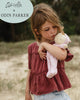 A young girl with tousled hair tenderly hugs an Olli Ella x Odin Parker Dozy Dinkums - Blossom doll while standing outdoors, wearing a maroon ruffled top. The text on the image reads "Olli Ella x Concordia Co x Aire Blooms.