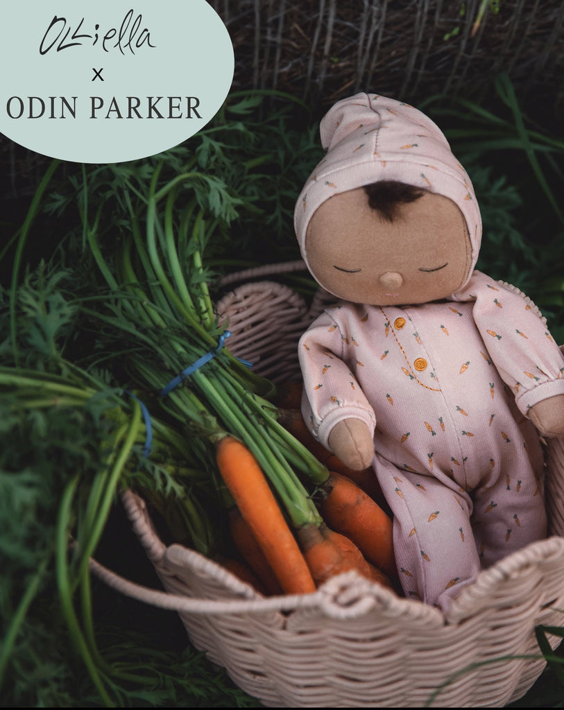 A Dozy Dinkum - Bugsy Hopscotch doll in a pink onesie rests in a wicker basket alongside fresh carrots, set against a natural green backdrop. The logo of "Olli Ella x Odin Parker" appears at the top.