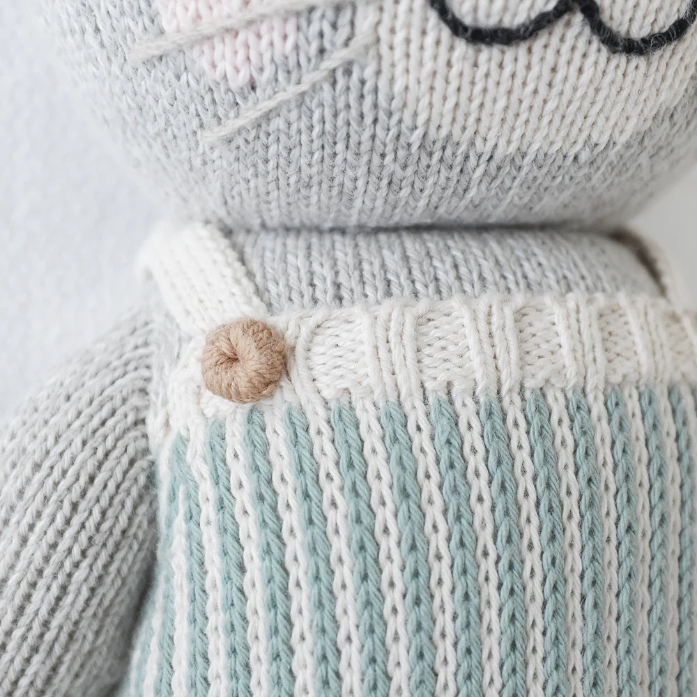 Close-up of Cuddle + Kind Dylan the Kitten, a hand-knit gray cat plush in a light blue and white striped outfit with a brown button on the strap; this stuffed toy features embroidered whiskers and mouth, perfect for snuggles.