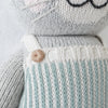 Close-up of Cuddle + Kind Dylan the Kitten, a hand-knit gray cat plush in a light blue and white striped outfit with a brown button on the strap; this stuffed toy features embroidered whiskers and mouth, perfect for snuggles.