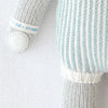 A close-up of the Cuddle + Kind Dylan the Kitten hand-knit doll, featuring grey and white arms, a white hand, and a blue and white vertically striped body on a soft, textured white background.