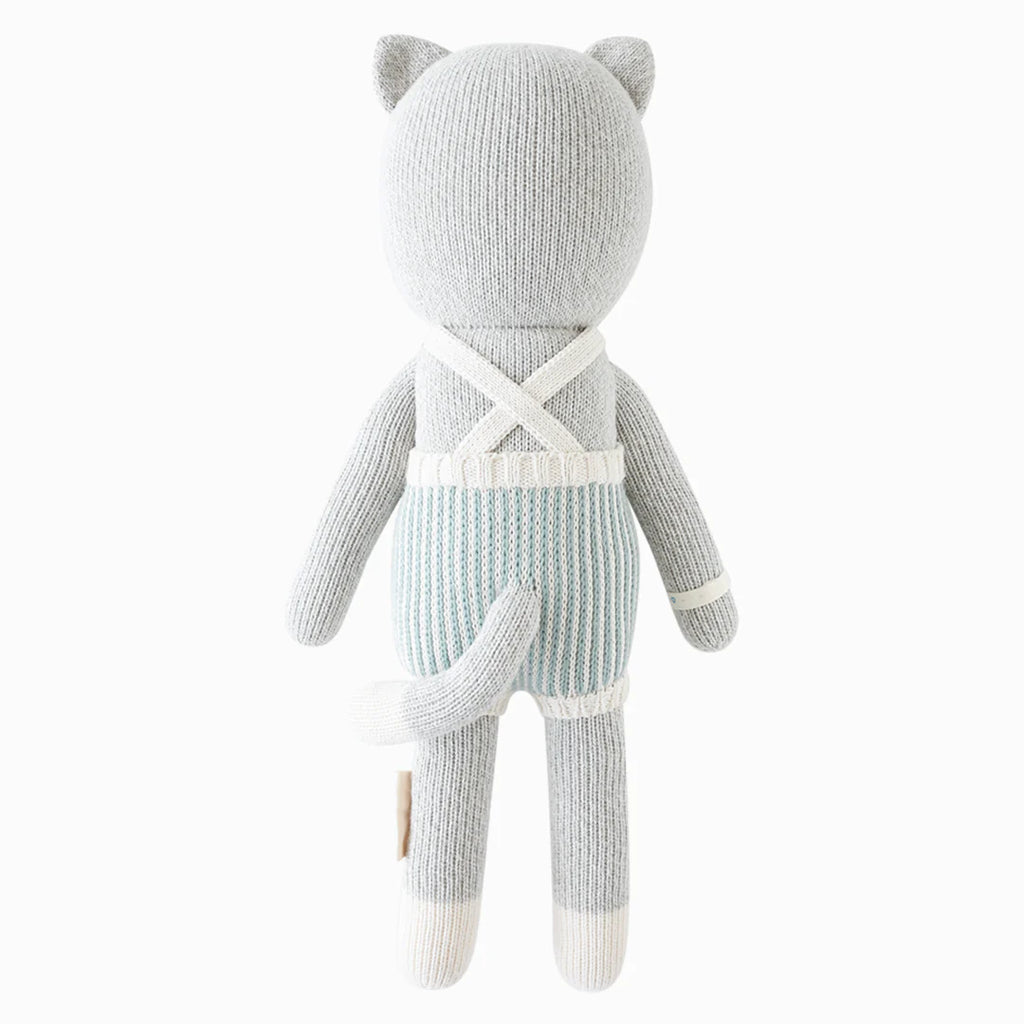Cuddle + Kind Dylan the Kitten is a hand-knit gray stuffed cat shown from the back, wearing blue and white striped overalls with white suspenders, a tail, round ears, and white accents on its hands, feet, and suspenders.