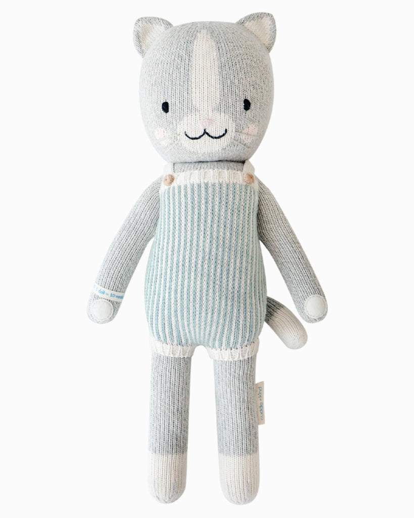 Cuddle + Kind Dylan the Kitten is a soft, hand-knit gray cat doll with white accents, a smiling face, and light blue striped overalls, featuring a simple and cute design. Shown on a white background.