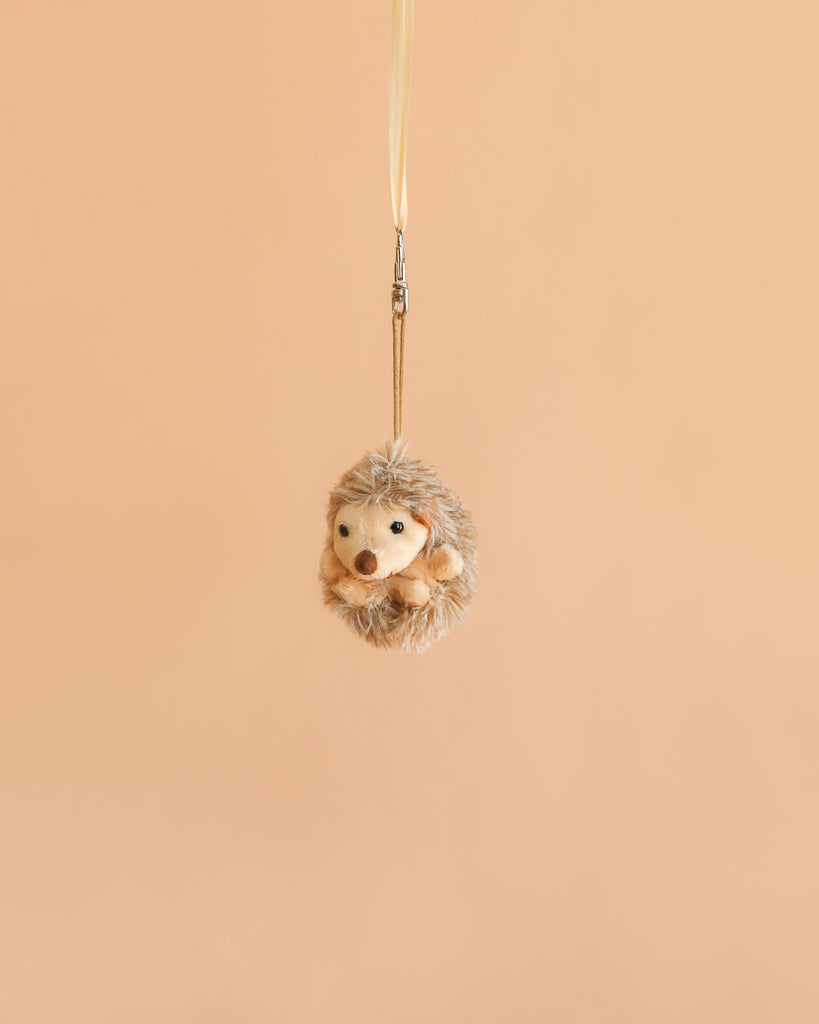 The Bukowski - Hubert Hedgehog Bag Charm, a small, round plush keychain, hangs from a thin strap against a plain, light beige background—a charming backpack accessory for any plush toy lover.