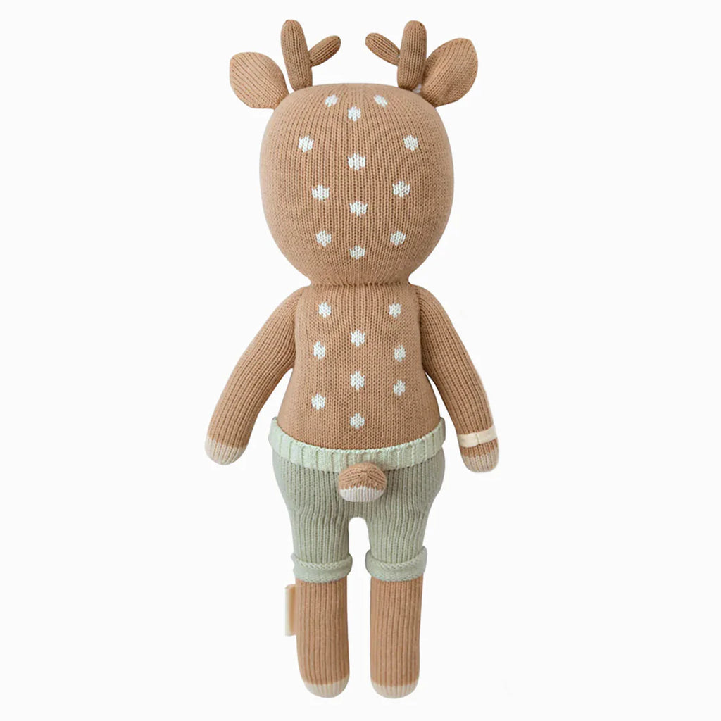 Cuddle + Kind Elliott the Fawn is a hand-knit, fair trade plush deer with light brown fur, white spots, antlers, and mint green pants. This hypoallergenic stuffed toy is shown from the back against a white background.